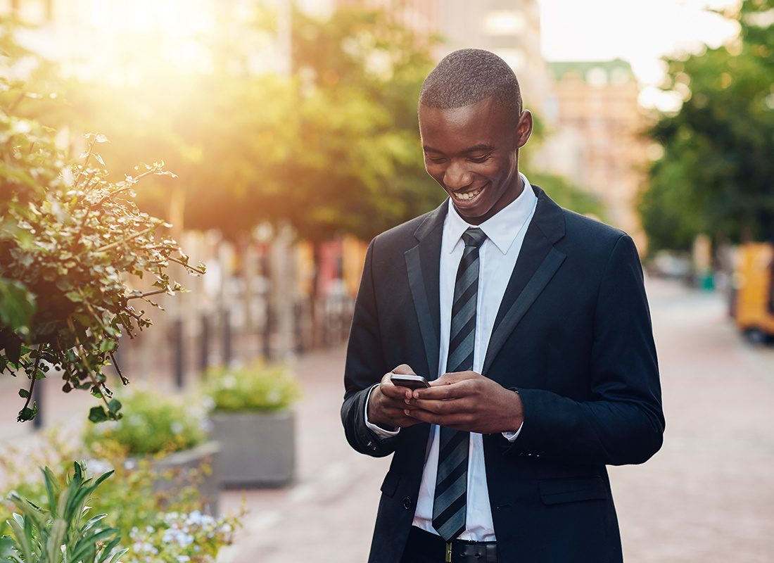 Homepage - Businessman Using a Smartphone and Smiling Broadly, Outside Surrounded by Trees and City Walking Paths
