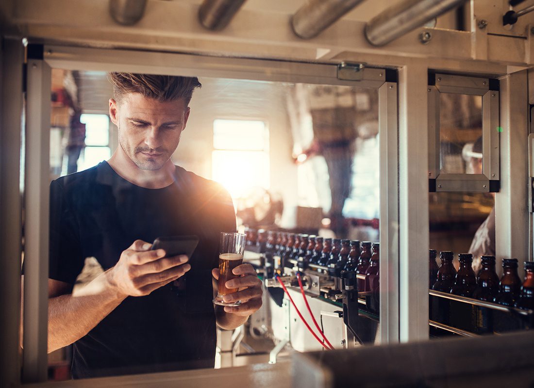 Request a Quote - Brewery Owner Using a Smartphone Surrounded by Equipment